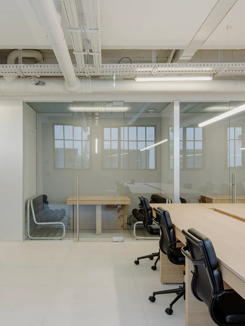 Open workspace with glass-enclosed meeting room, light wood desks, and exposed white structural beams overhead