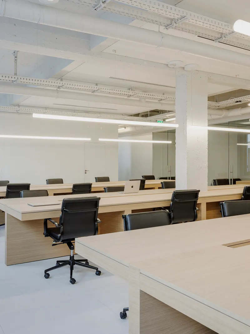 Rows of light wood desks with black office chairs under painted white structural beams and ductwork