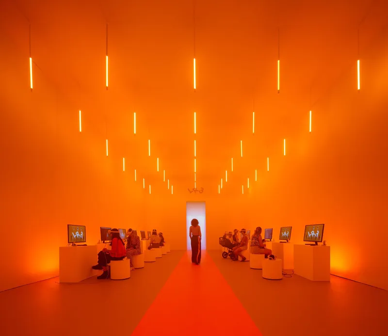 Gallery interior bathed in orange light with suspended linear fixtures and visitors seated on cylindrical stools
