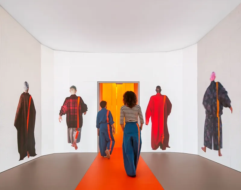 White curved gallery space with orange floor path leading visitors toward a glowing orange doorway