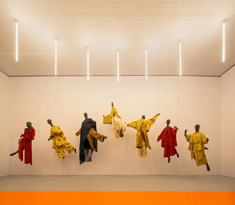 Exhibition wall with suspended garments in yellow and red under linear ceiling lights and orange floor stripe