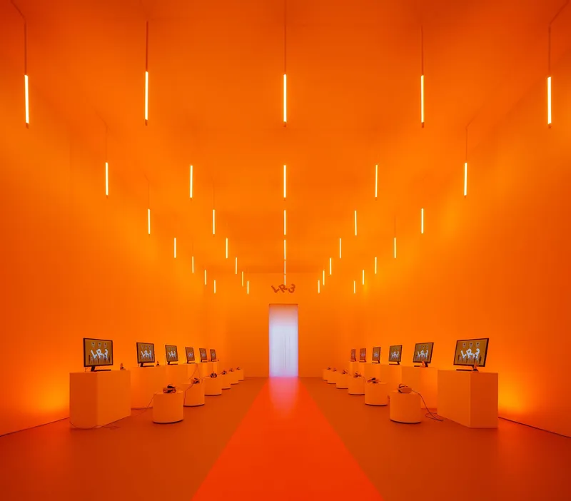 Gallery space with orange walls, vertical light tubes suspended from ceiling, and low display plinths