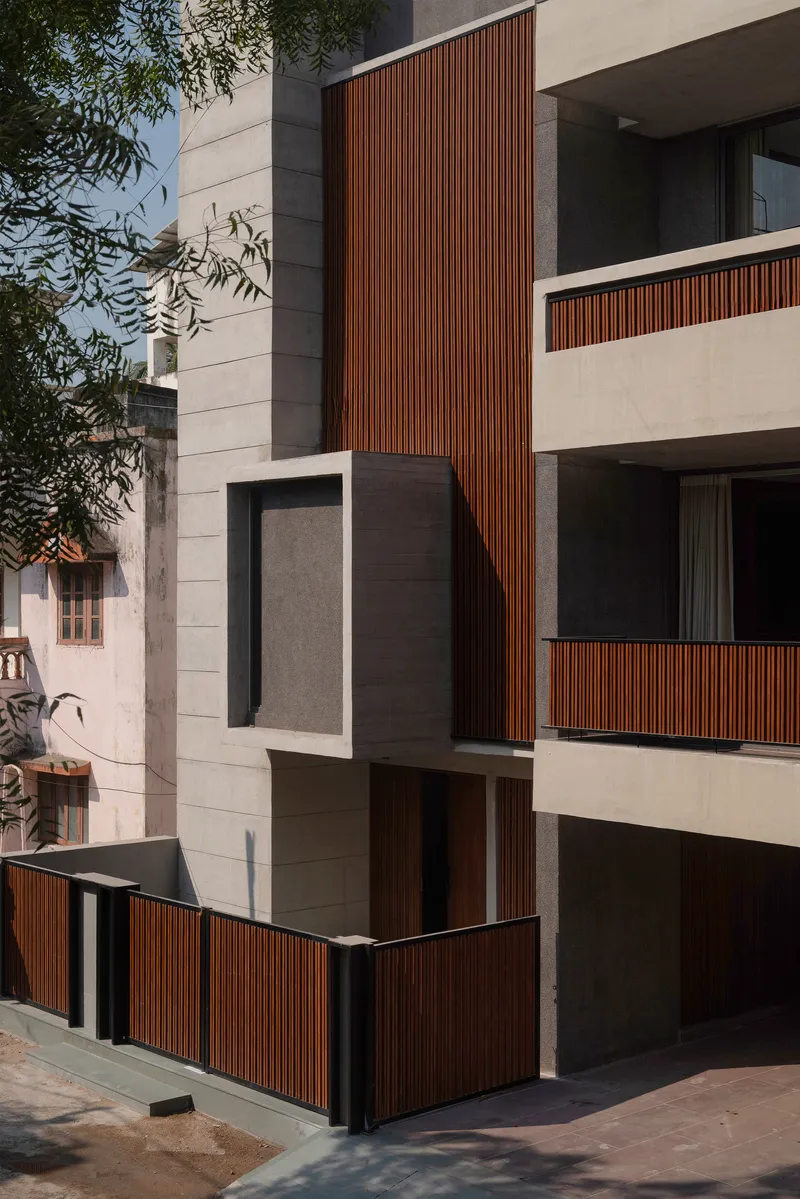 Corner entrance featuring vertical timber slats and stacked concrete balconies framed by overhanging tree branches