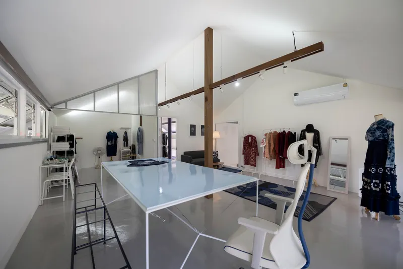 Interior workspace with white worktable, exposed timber beams crossing overhead, and garments displayed on wall