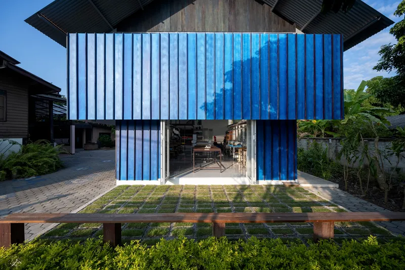 Blue corrugated metal volume with folding glass doors opening onto a gridded paver and grass courtyard