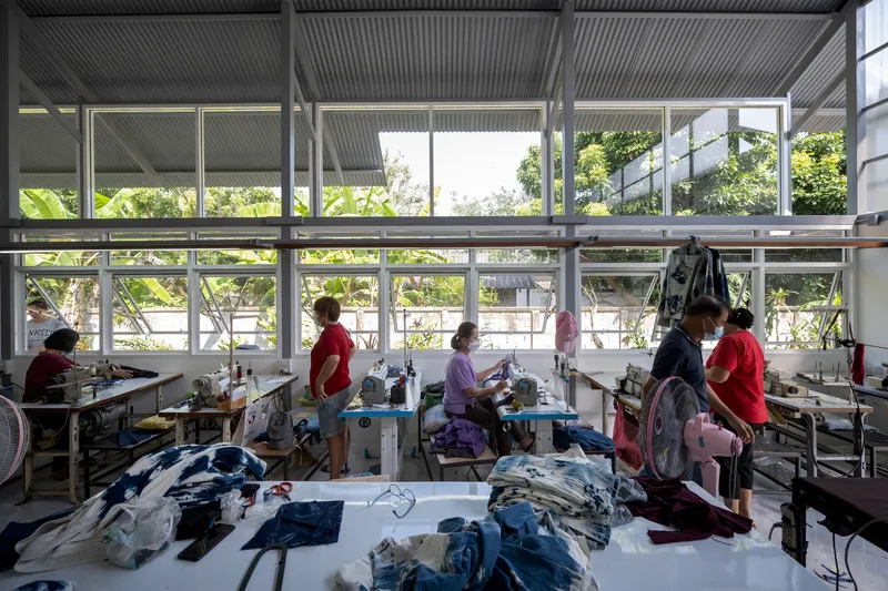 Workshop interior with workers at sewing stations beneath steel frame windows overlooking planted vegetation