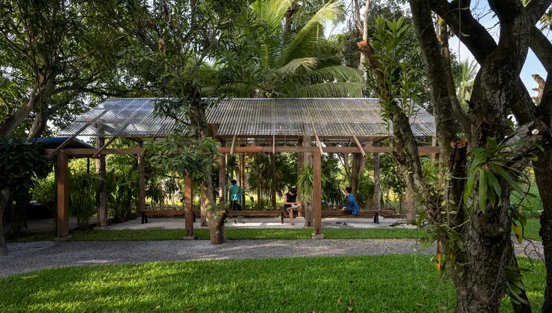Open-sided timber pavilion with corrugated metal roof sheltering a workspace among trees and tropical plants