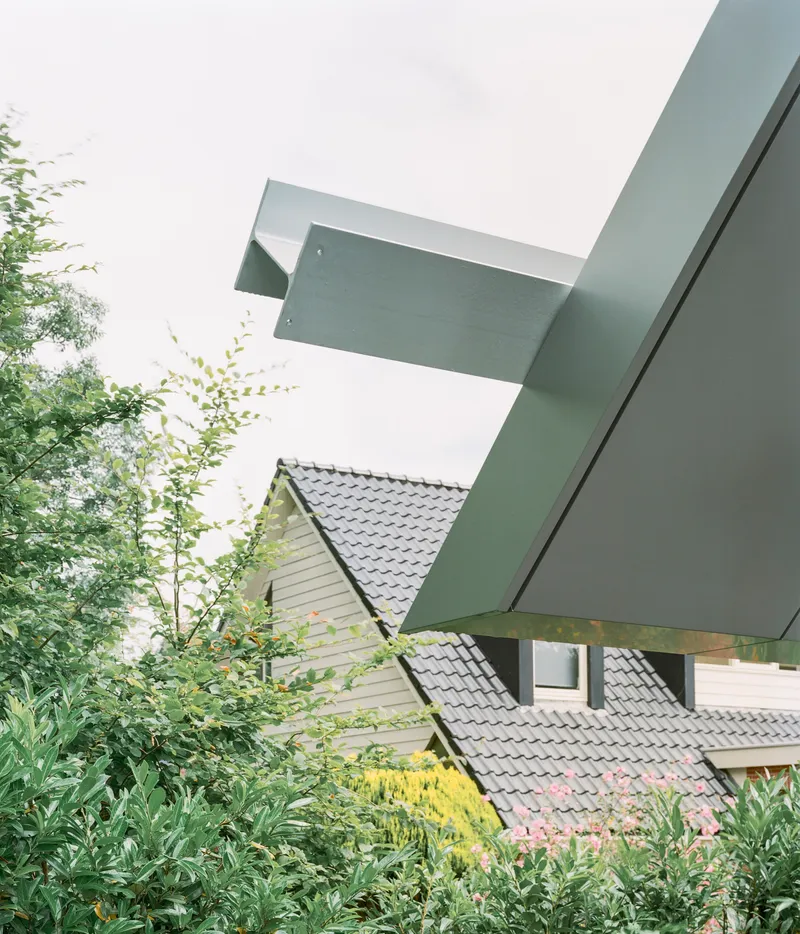 Cantilevered metal roof planes extending over neighboring tiled roof and garden plantings