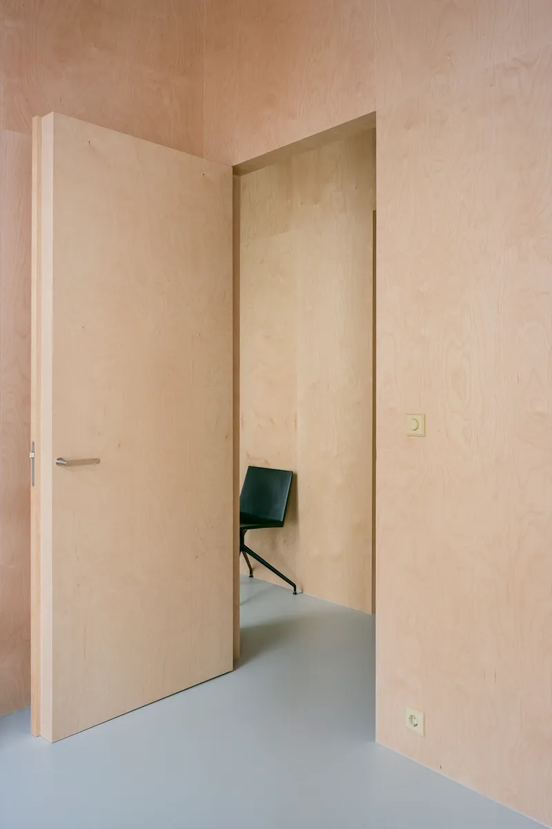 Pale plywood-lined hallway with flush door and glimpse of black chair beyond