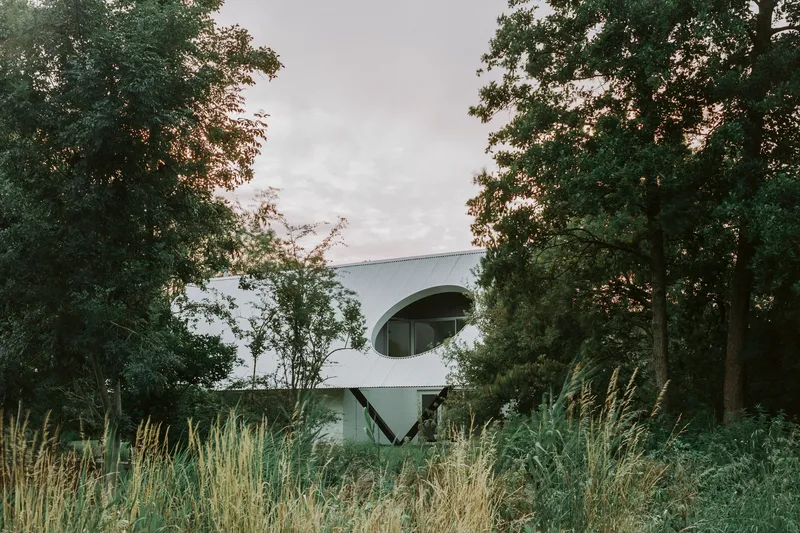 White corrugated metal facade with circular opening framed by tall trees and wild grasses at dusk