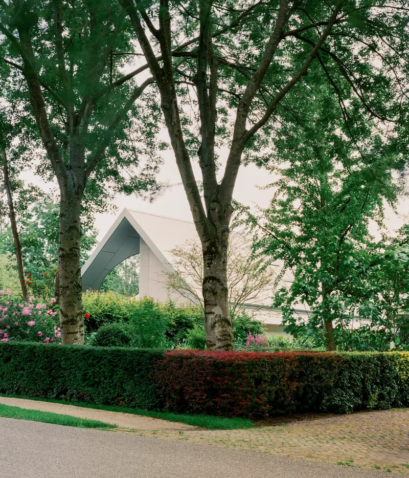 White gabled roof viewed through mature trees and trimmed hedges along a curved path