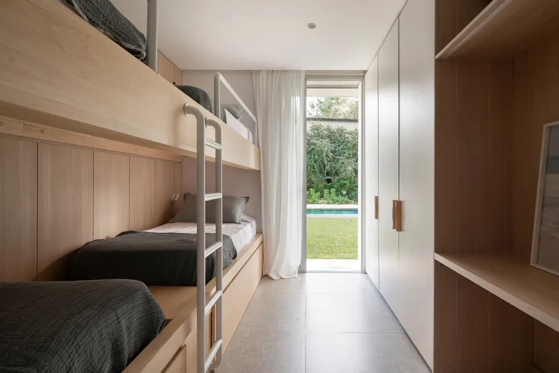 Bunk bed room with blonde timber casework and white curtains framing a view to lawn and pool