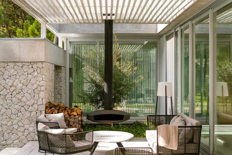 Covered terrace with suspended black fireplace, stone mosaic wall and slatted pergola filtering daylight