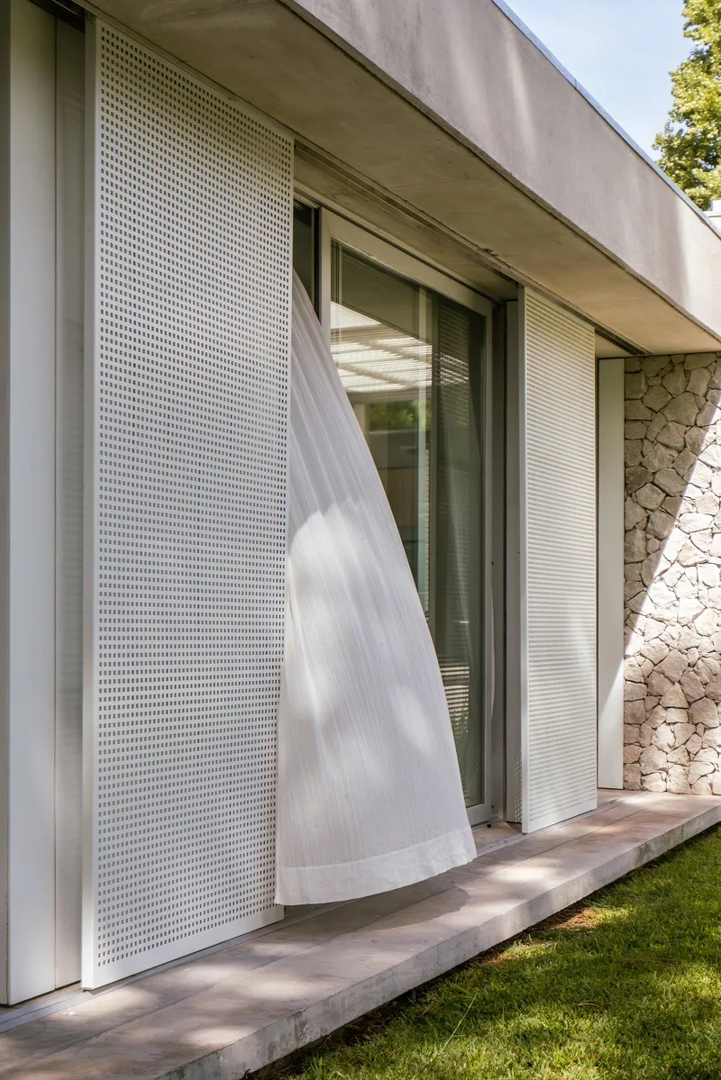 Exterior concrete overhang with perforated metal screen and billowing sheer curtain beside stone wall