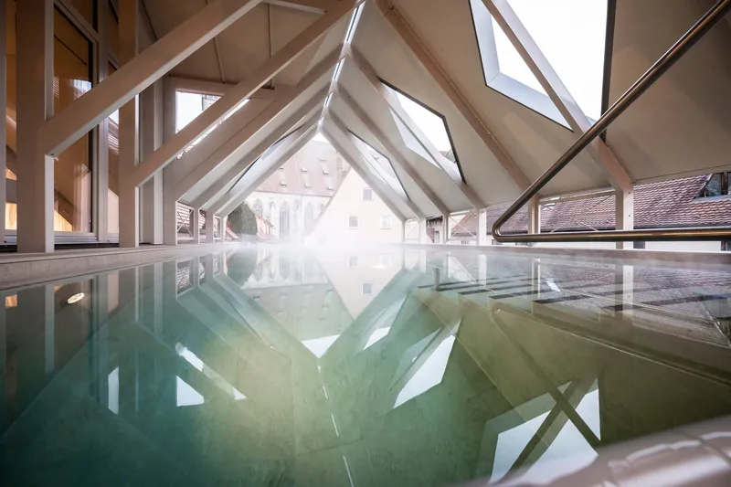 Indoor pool beneath a timber-framed gable roof with angled glazing reflecting the surrounding buildings