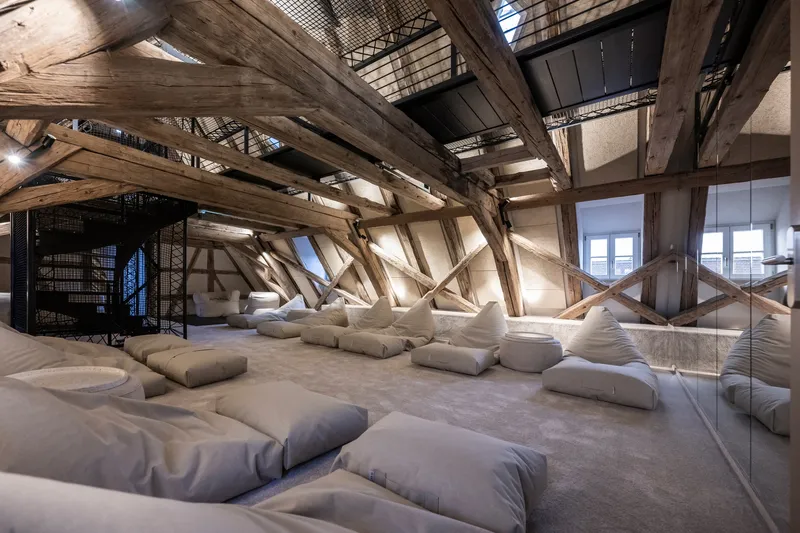 Attic relaxation room with exposed diagonal timber beams and floor cushions scattered on polished concrete
