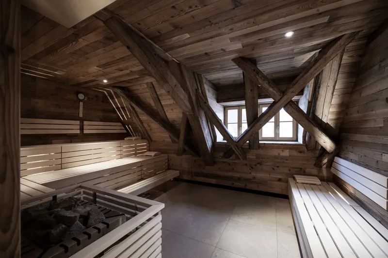 Timber sauna interior with diagonal bracing beams, tiered benches, and a corner stove heater