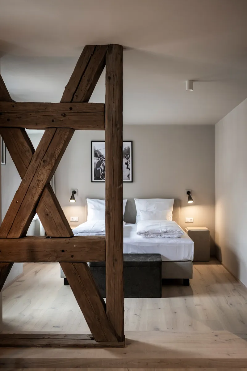Exposed timber frame with diagonal cross bracing dividing a bedroom with white linens and wall sconces