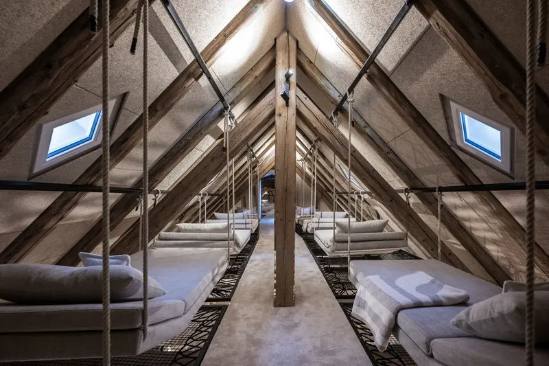 Attic dormitory with twin rows of suspended bunk beds under exposed timber roof trusses and skylights