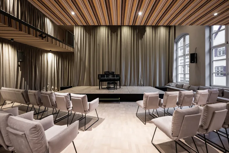 Stage platform with grand piano facing semicircular seating arrangement beneath fabric curtains and slatted acoustic ceiling