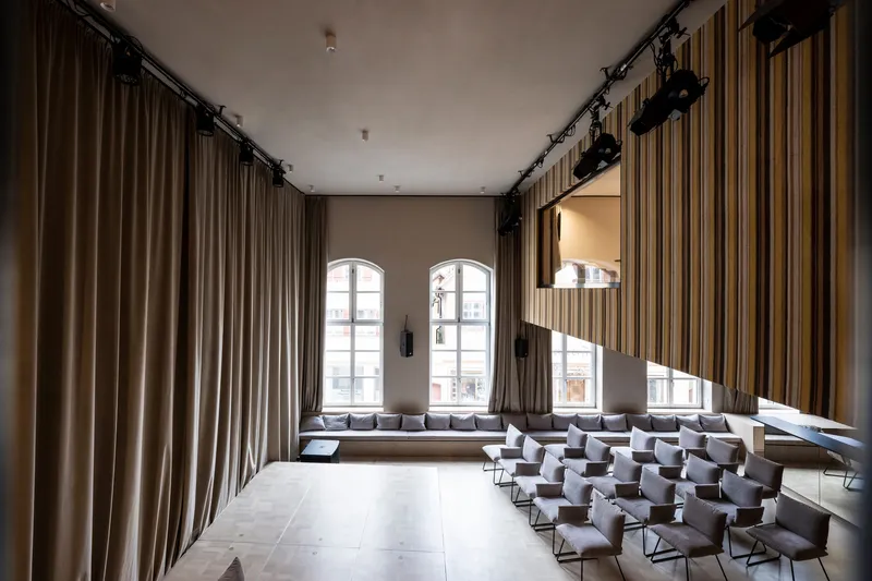Double-height performance space with built-in window benches and angled sunlight across the pale wood floor