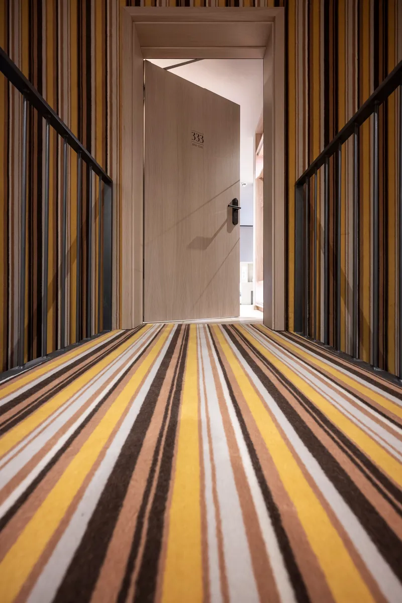 Corridor with striped carpet in yellow and brown tones leading to a numbered door framed by vertical wood slats