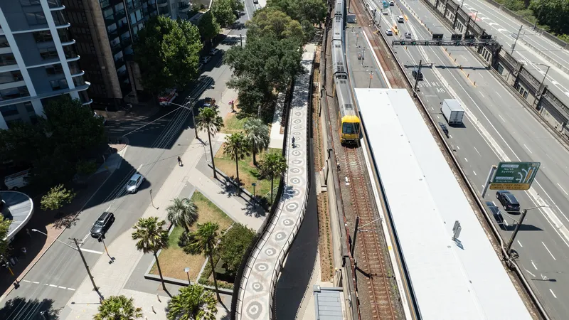 Overhead view of the elevated railway platform with white canopy roof running between landscaped pathways and highway