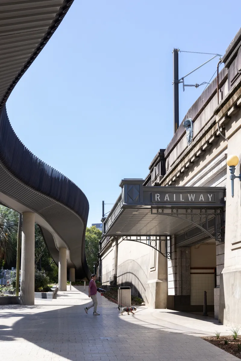 Curved elevated walkway with corrugated metal balustrade meeting the heritage railway station entrance below