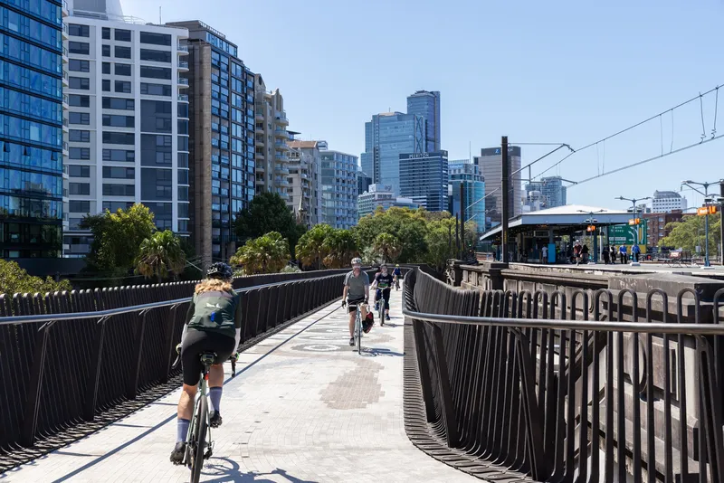 Cyclists and pedestrians on the undulating bridge deck with vertical metal balusters and urban skyline beyond