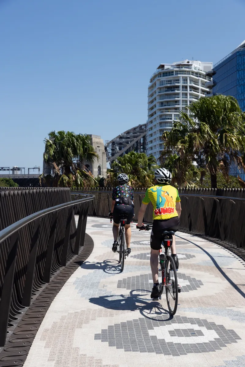 Elevated pathway with dark vertical balustrade and cyclists riding toward residential towers in sunlight