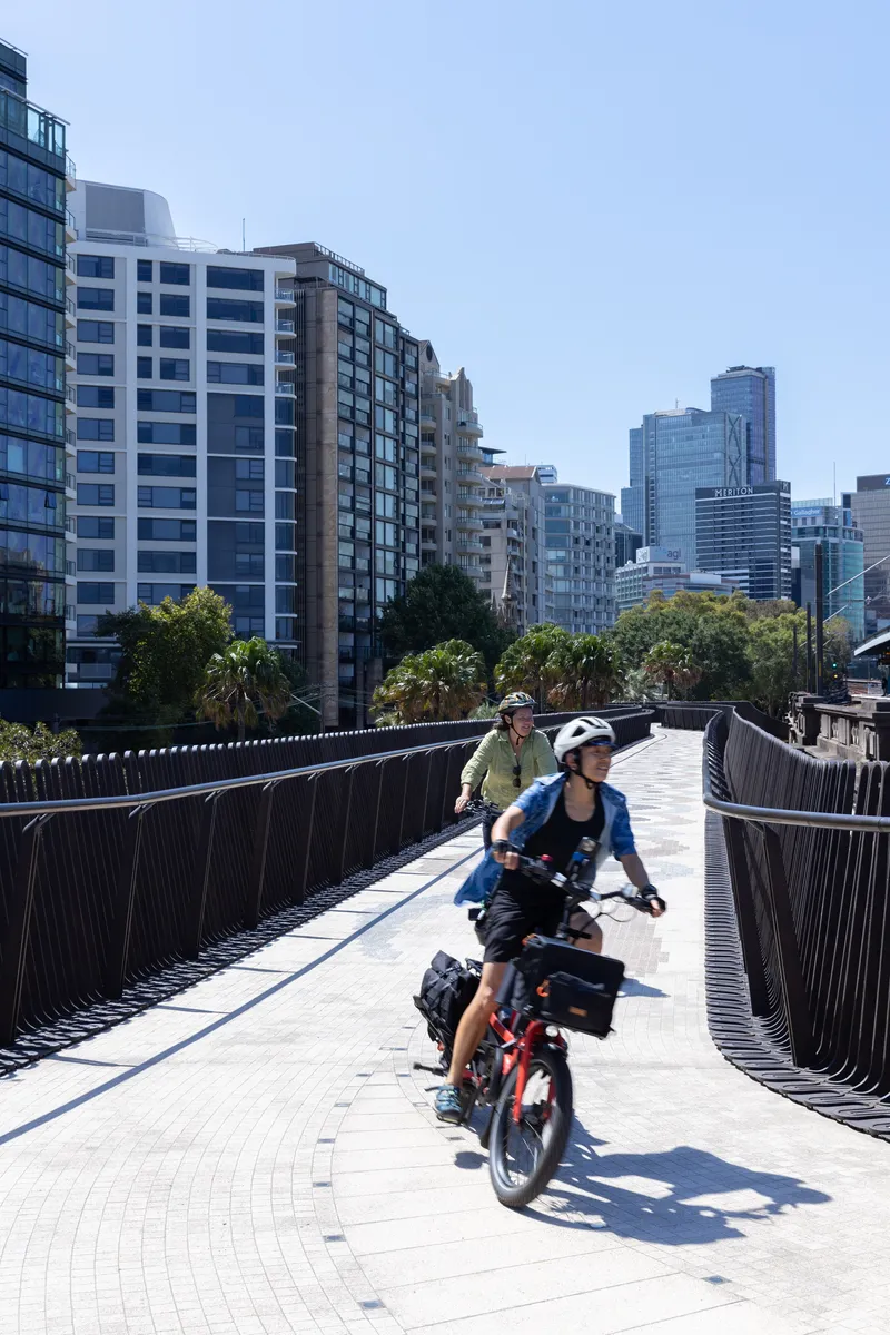 Curved cycling path bordered by fluted metal barriers with riders heading toward urban skyline