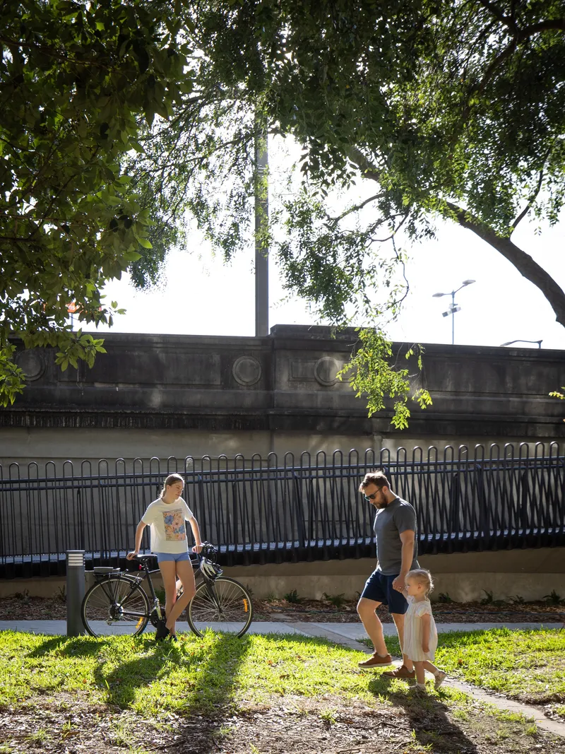 Metal railing along heritage viaduct wall with family and bicycles in dappled shade below trees