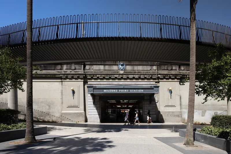 Station entrance beneath curved pedestrian bridge with vertical metal railing and people walking through archway