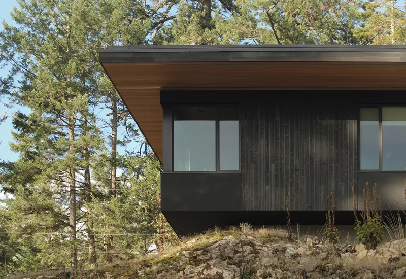 Cantilevered roof with cedar soffit extending over charred wood cladding and hillside boulders