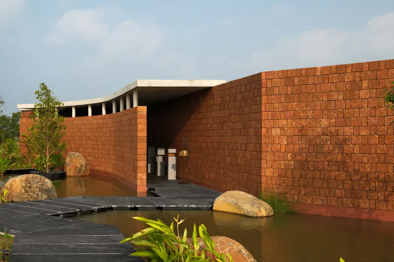 Laterite brick entrance wall with dark timber boardwalk crossing a reflecting pool flanked by boulders and plants