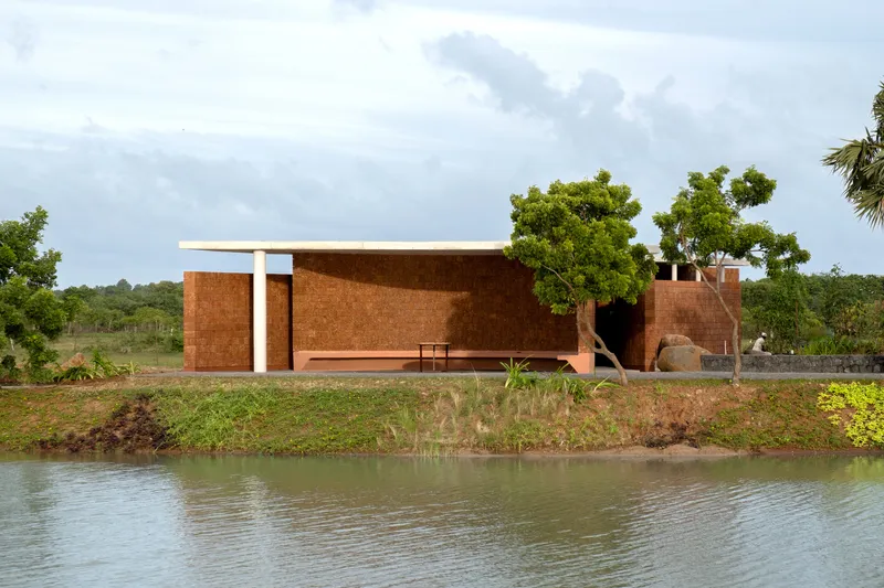 Long laterite brick wall with concrete overhang and recessed openings viewed across a pond with grassy banks
