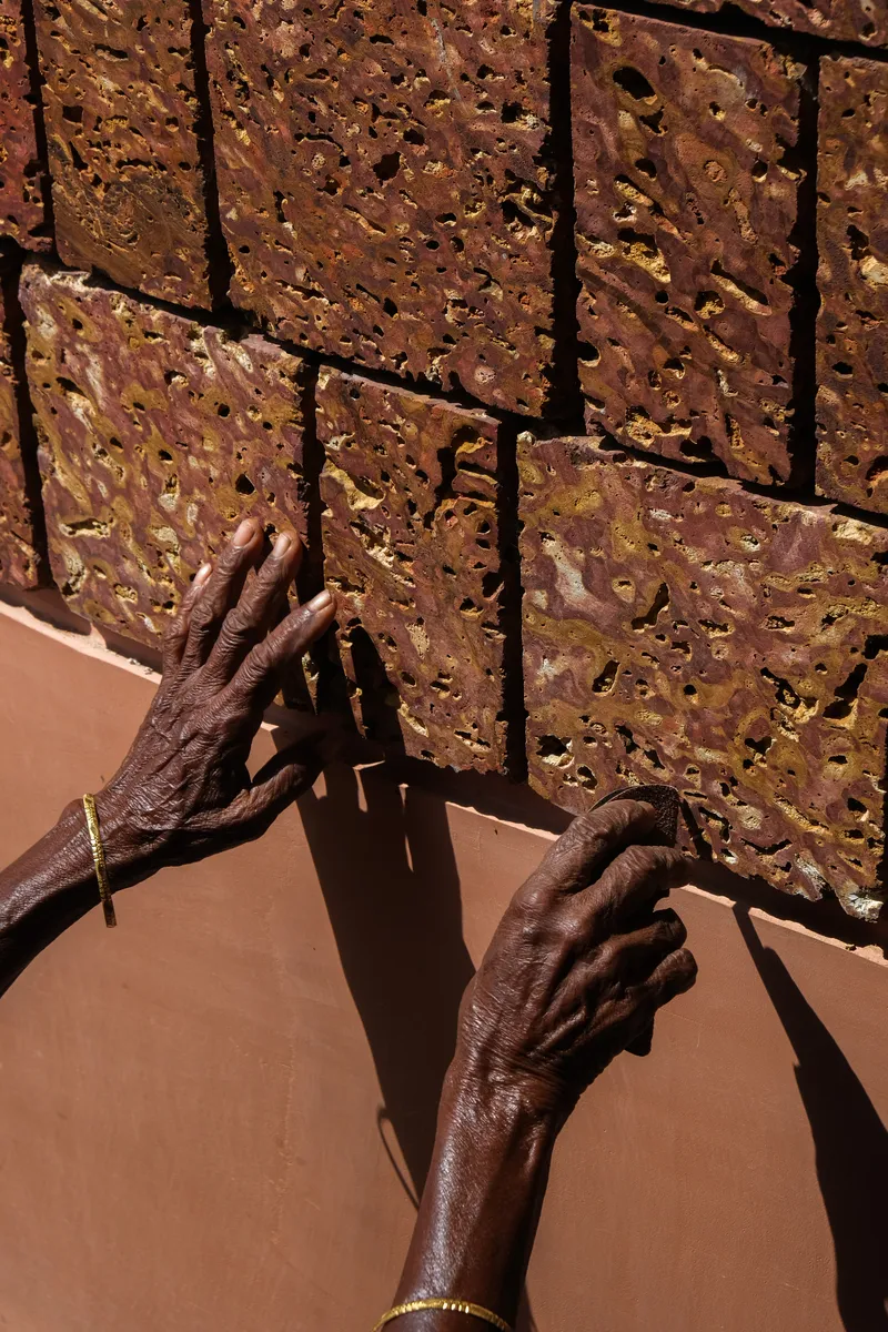 Close-up of hands touching textured laterite brick wall showing irregular surface and deep mortar joints in sunlight