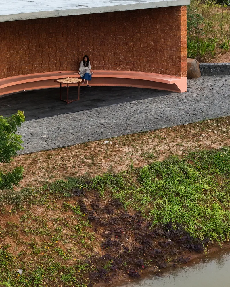 Curved terracotta bench against laterite brick wall with seated figure on dark stone paving beside pond