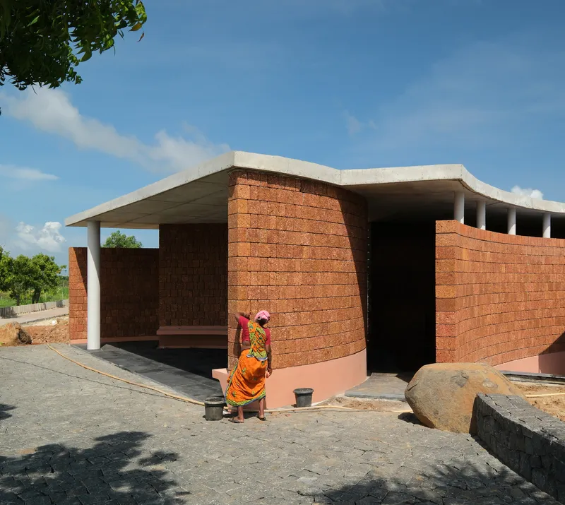 Exterior view of curved compressed earth block walls beneath a concrete roof slab as a woman walks past