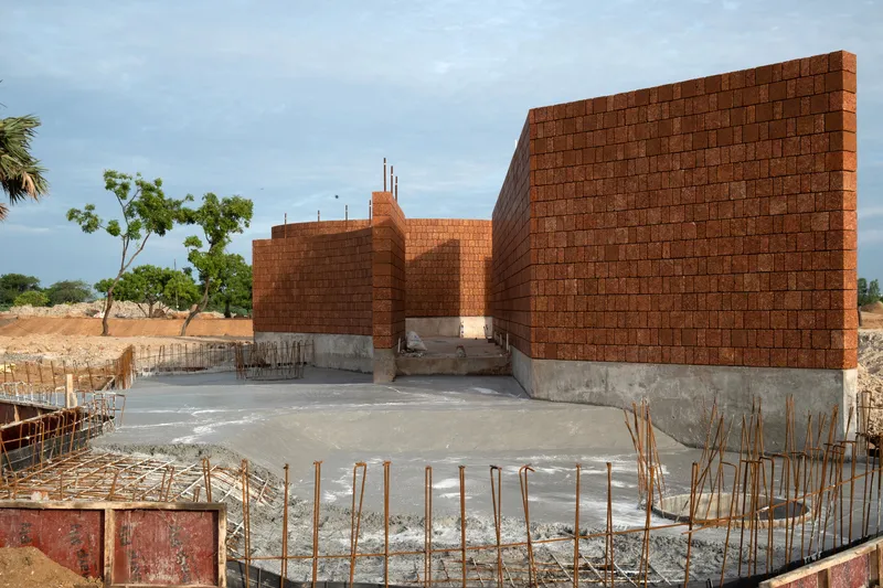 Construction site showing compressed earth block walls rising from concrete foundations with exposed rebar