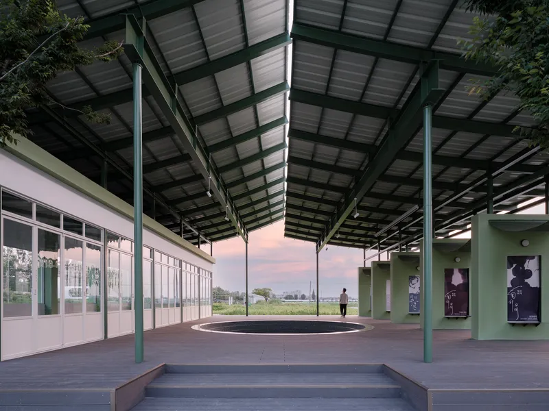 Covered outdoor platform with exposed steel structure, corrugated metal roof, and glazed classroom volume at dusk