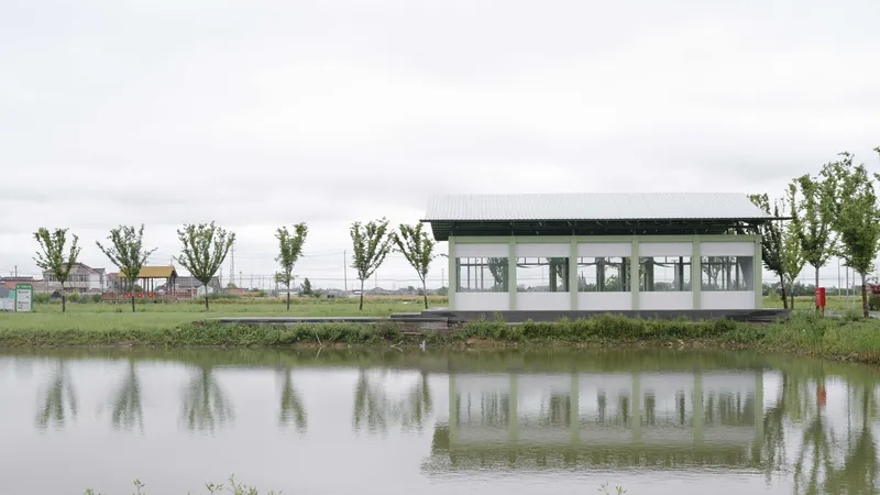 Glazed pavilion reflected in the still pond under overcast sky with row of young trees
