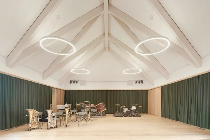 Rehearsal hall with exposed timber roof trusses, circular pendant lights, and green acoustic curtains surrounding brass instruments