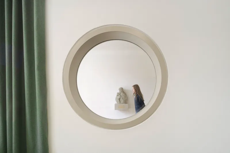 Circular window opening framing a visitor and a sculpture inside with green curtains visible at left