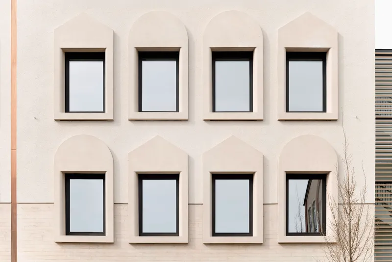 Facade detail of arched window openings with dark frames set in pale stucco beside a bare tree