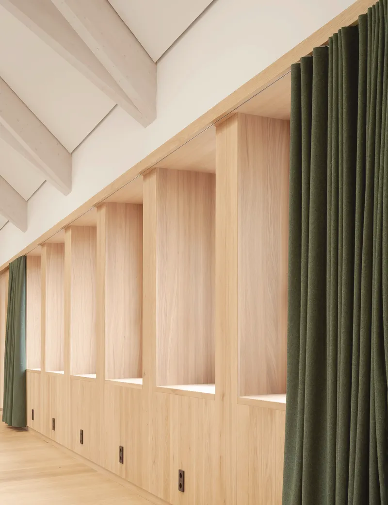 Light wood storage wall with integrated benches and olive green curtains beneath a sloped ceiling