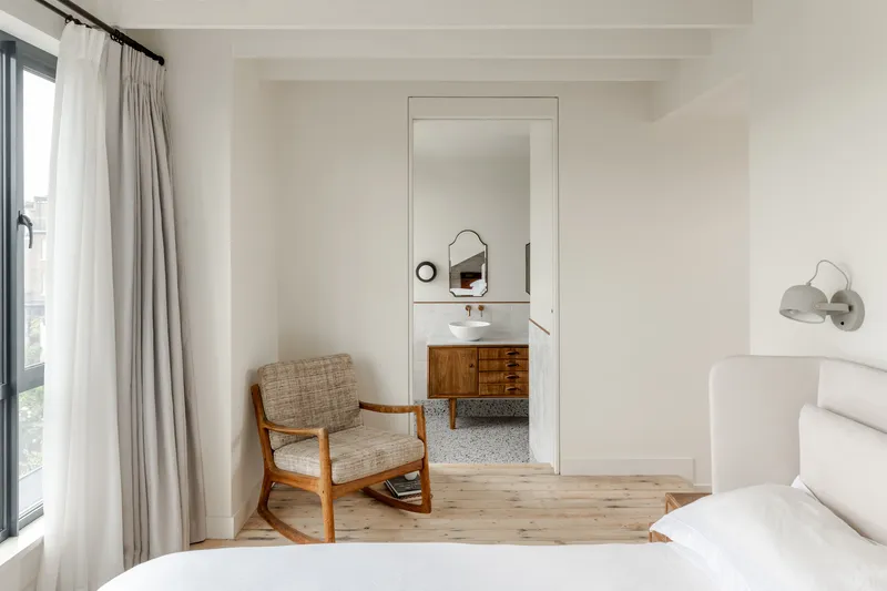 Bedroom with timber rocking chair looking through doorway to ensuite bathroom with terrazzo flooring