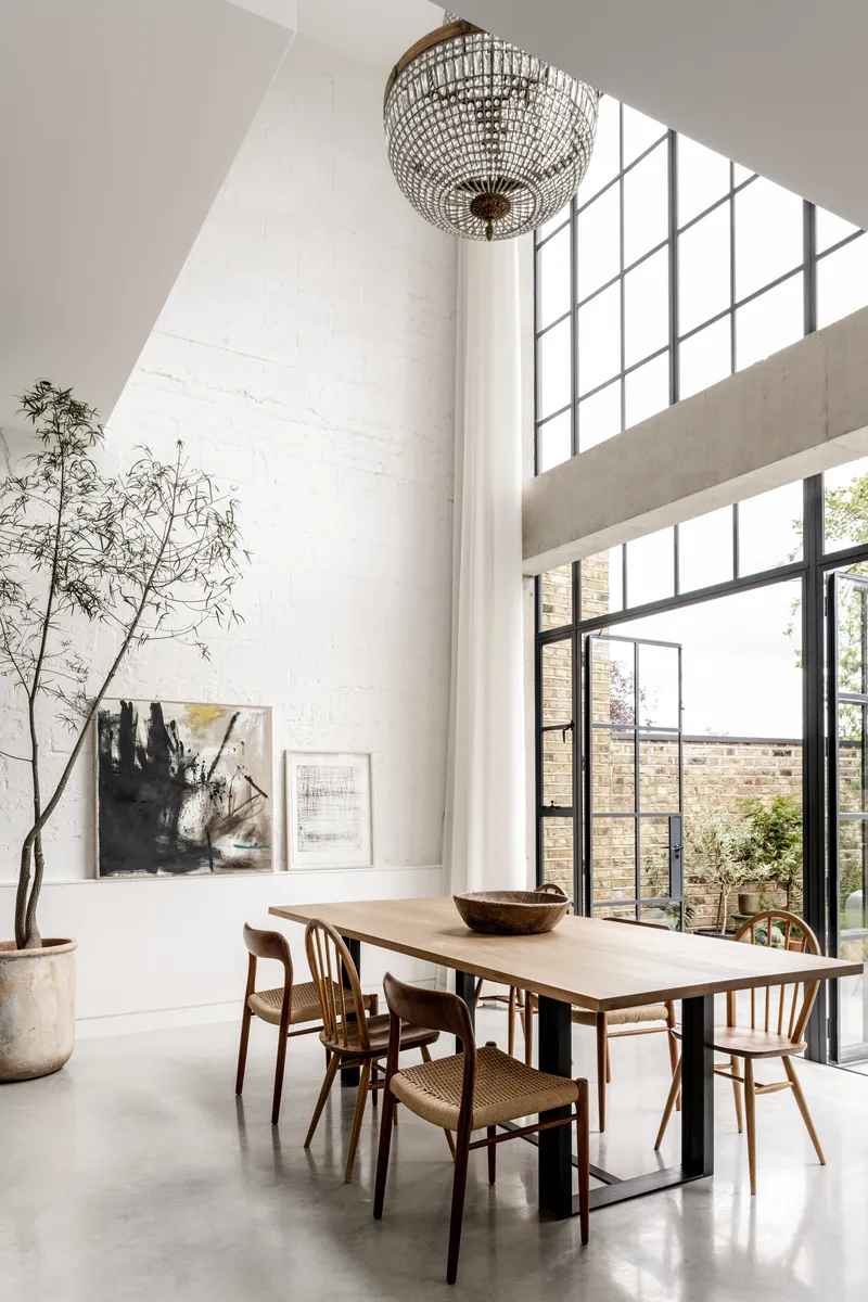 Double-height dining room with crystal chandelier and tall steel-framed windows framing courtyard trees