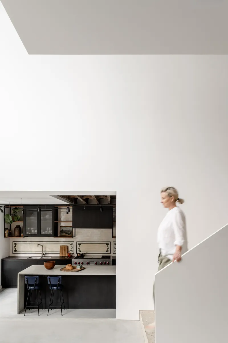 Person ascending a sculptural white staircase beside the black kitchen with exposed timber ceiling