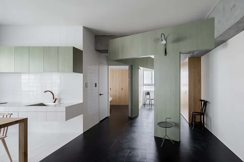 Open-plan interior with white kitchen island, pale green cabinetry, and black flooring under exposed concrete beams
